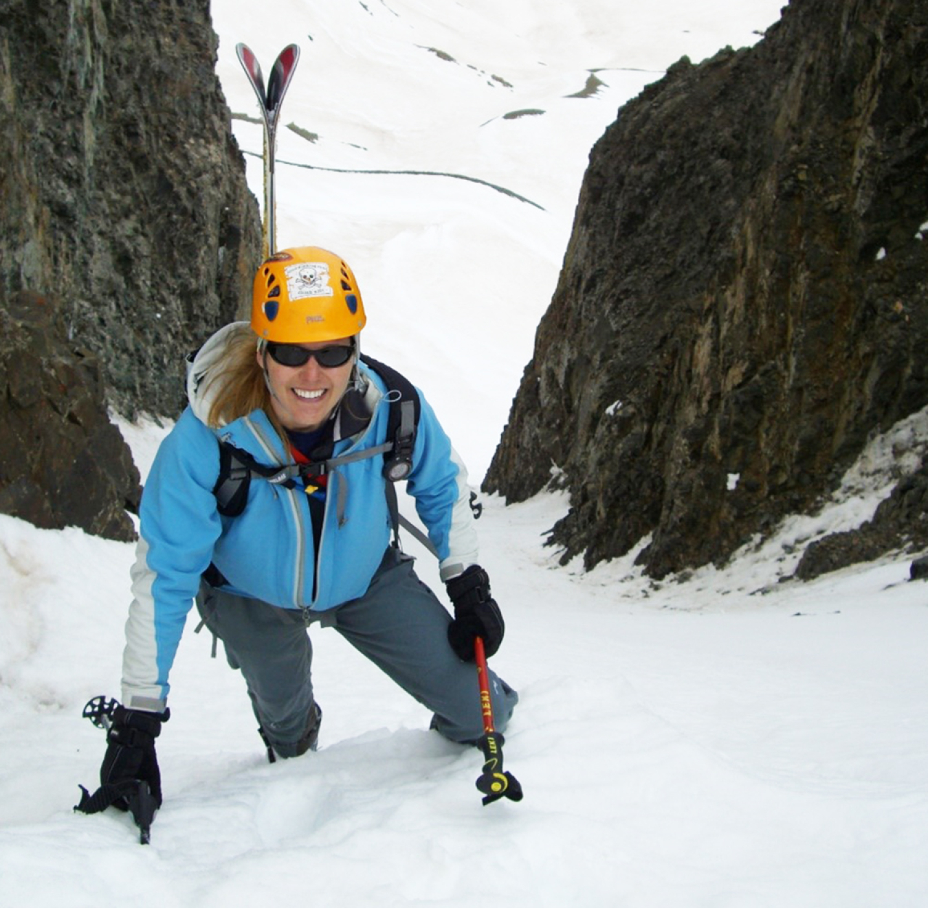 Amy Bauer dressed in skiing gear, poses on a steep slope of snow. She is holding downhill ski poles in her hand and smiling. Her skis are stuck in the snow behind her.