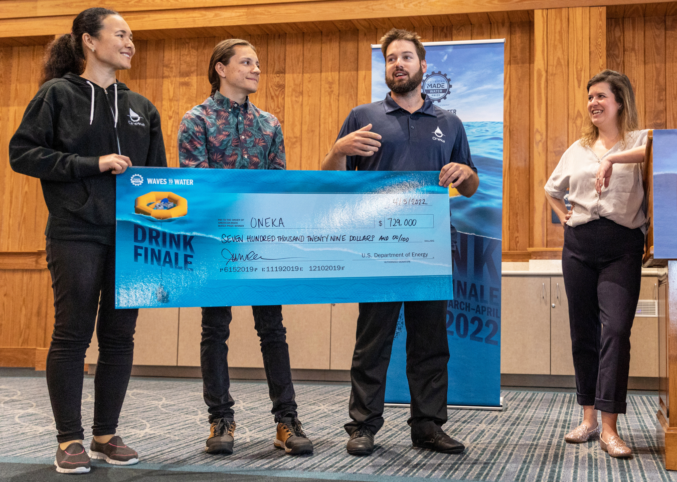 Three people holding a large check, and one person standing to the side next to a podium.