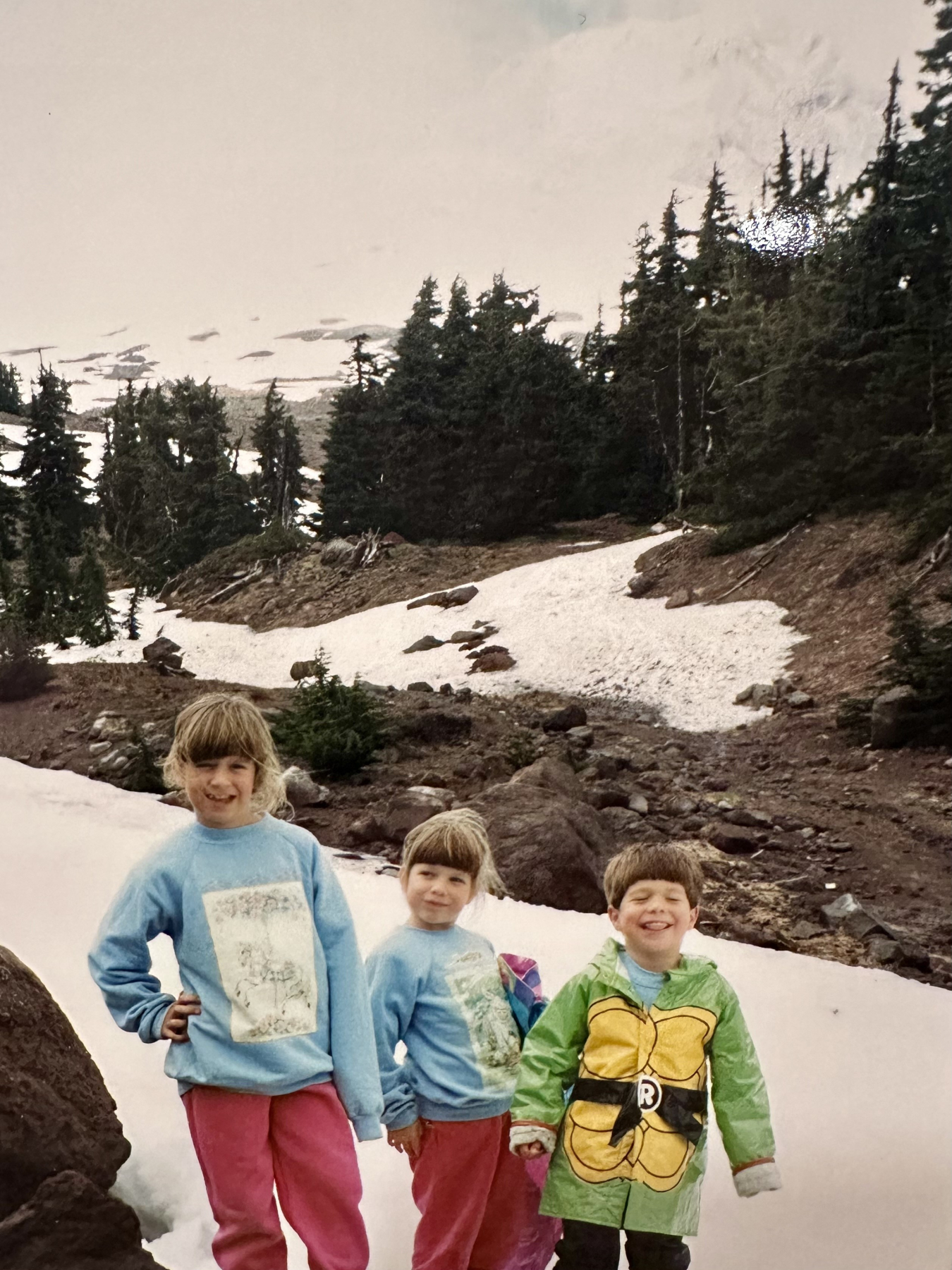 Three children posing for a picture with a rocky, somewhat snow-covered landscape behind them.