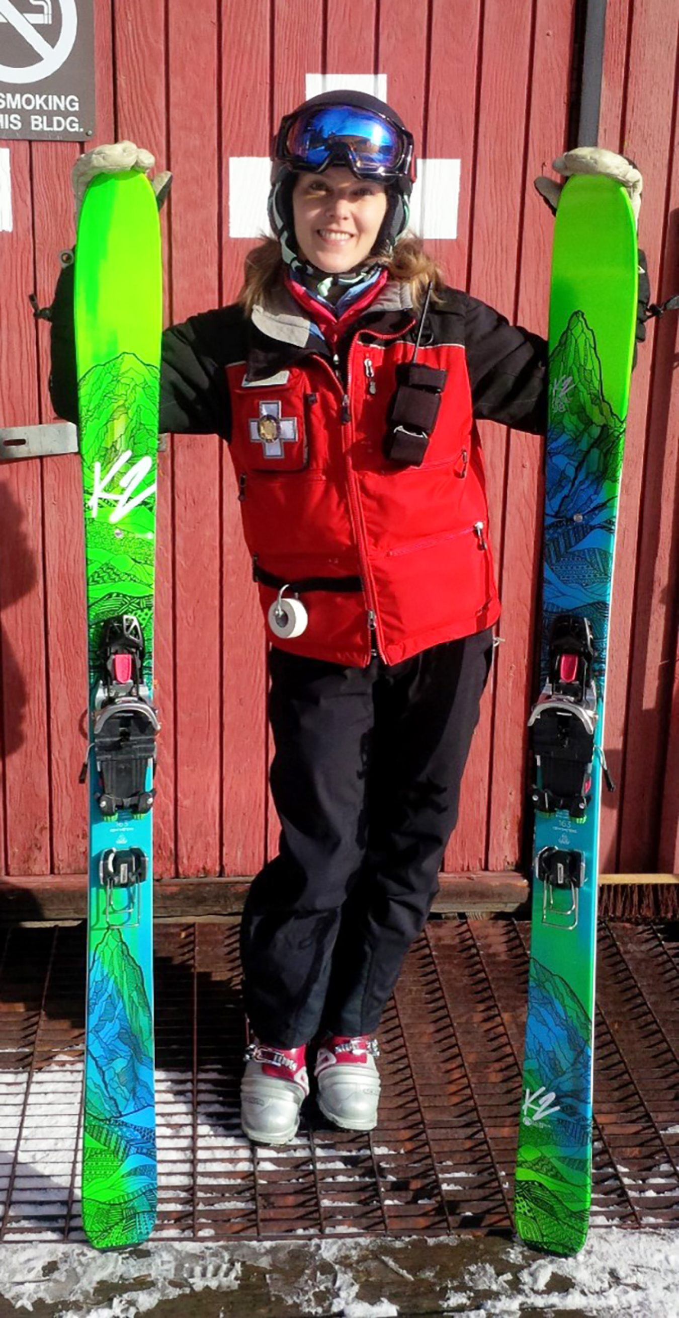 Amy Bauer poses with her downhill skis vertical on either side of her. She is dressed in a ski patrol outfit.