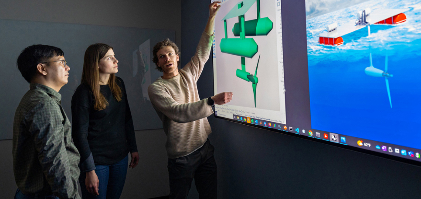 Three researchers looking at an enlarged computer screen featuring illustrations of two tidal energy devices. 