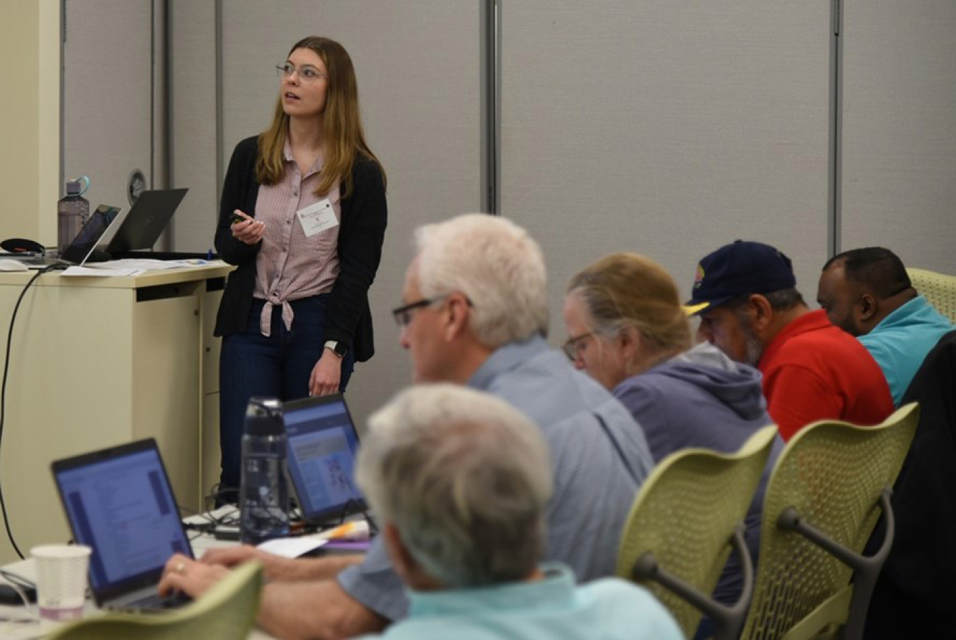 A woman speaking at a podium in front of a crowd, looking at her presentation (off screen).