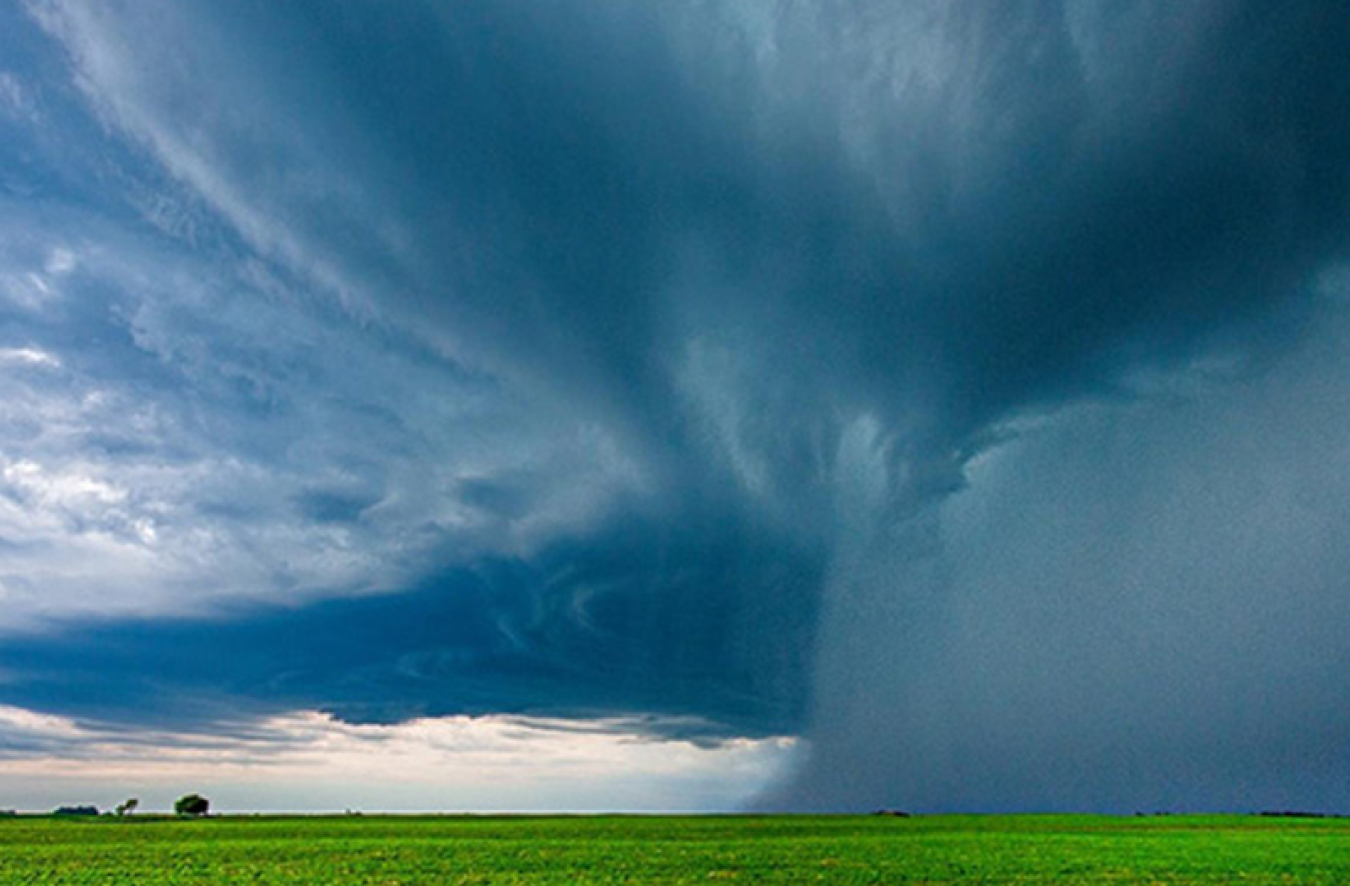 A huge, rolling, gray cloud over a field of green with a house very far in the background. 