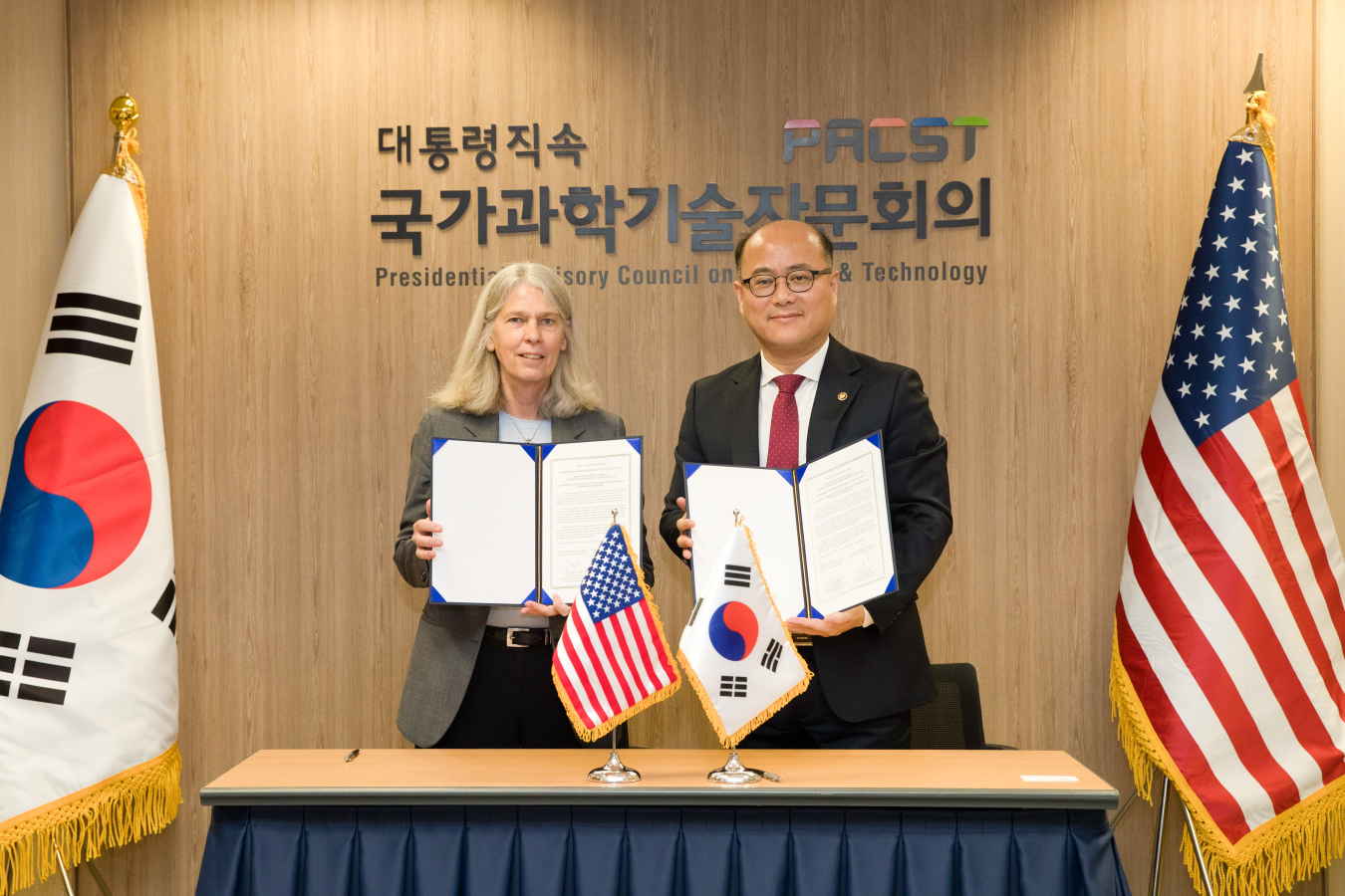 A photo of Jill Hruby and Lee Chang Yune holding up signed agreements. U.S. and Republic of Korea flags are to their sides. On the wall behind them, in English and Korean, it says "Presidential Advisory Committee on Science and Technology."