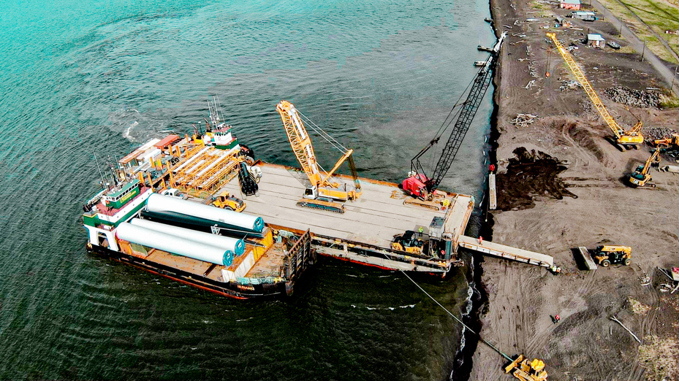 A barge with wind turbine tower and blade pieces being unloaded on to a floating dock by several small cranes.