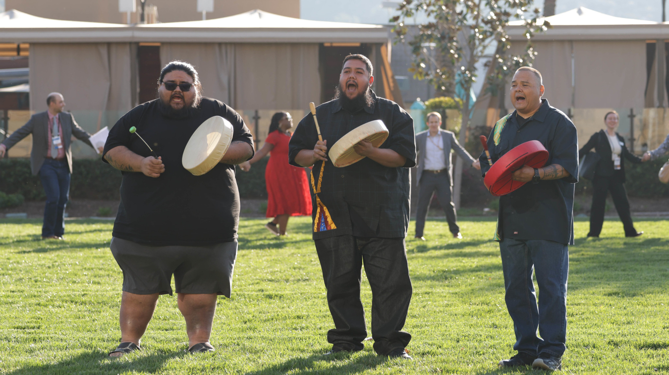 Three men with hand drums sing while a circle of adults in professional clothing holding hands dance behind them.