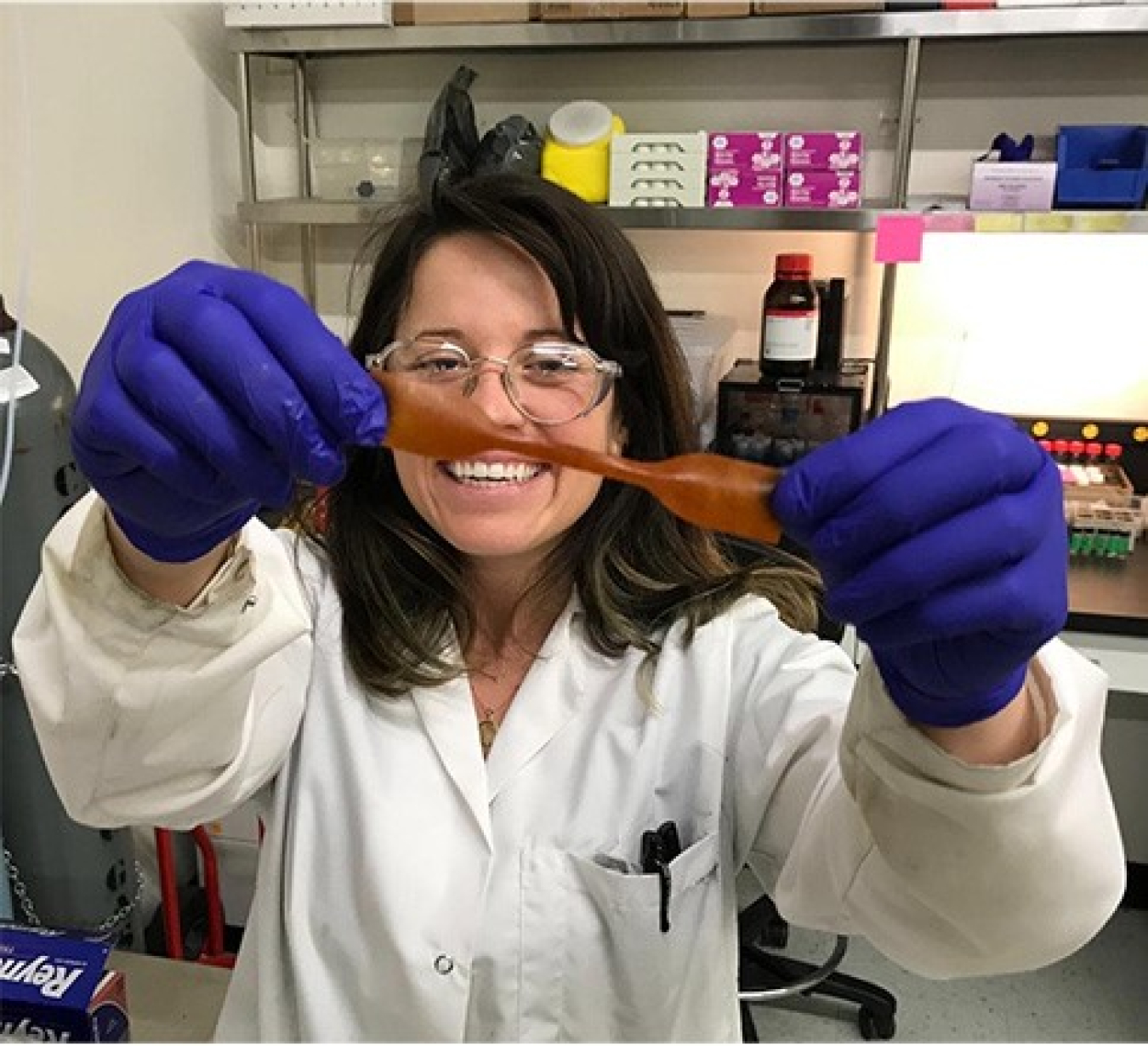 Smiling Kat in gloves, lab coat, and goggles, holding up a piece of materials toward the camera.