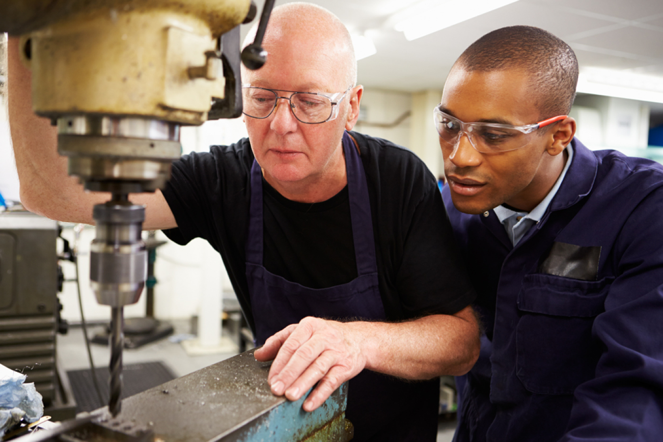 Two people, wearing safety equipment, work at a drill press