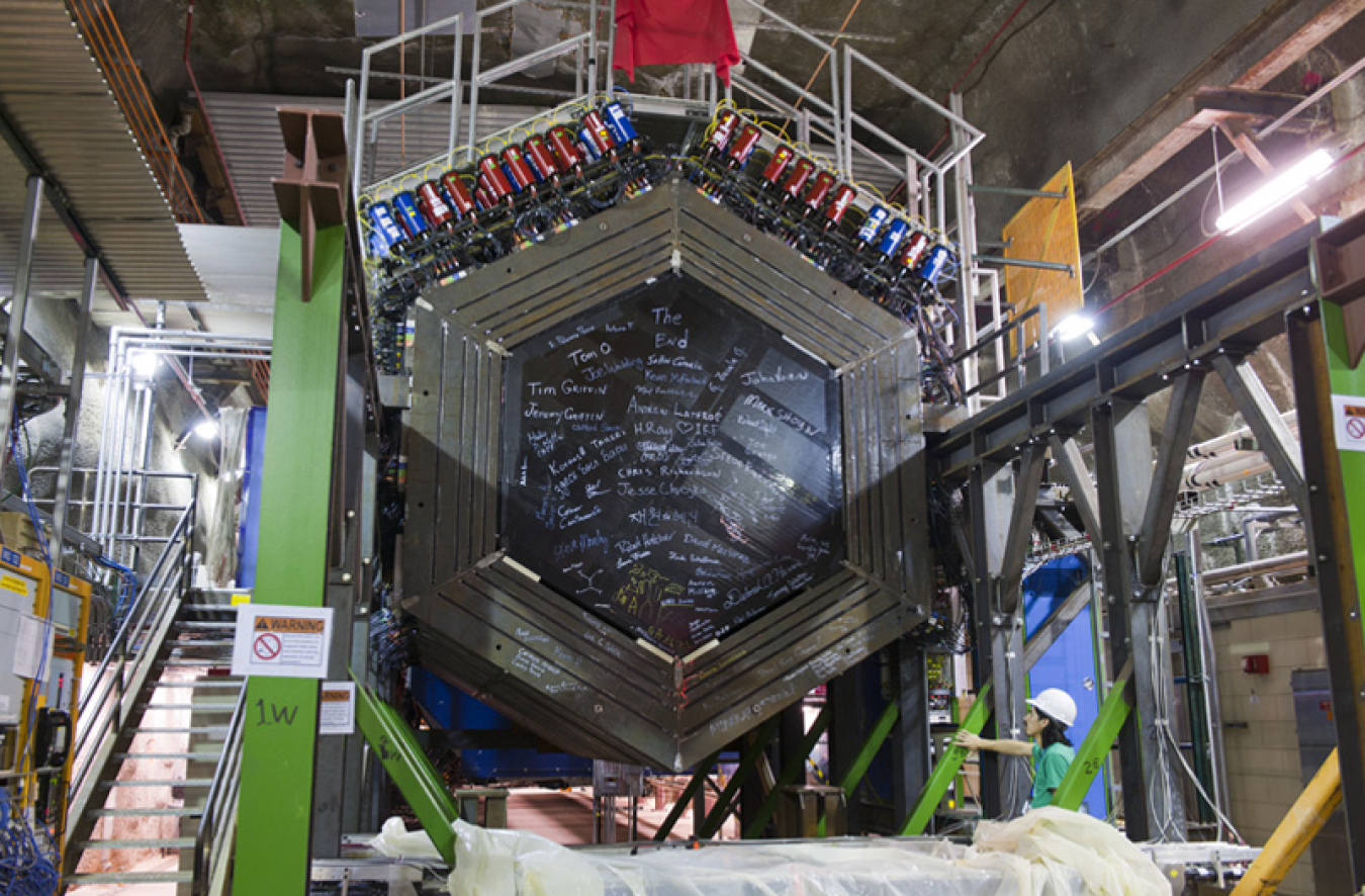 The particle detector used in the MINERvA experiment. It is a large hexagon with blue and red cylinders along the top and signatures decorating the face of the detector.