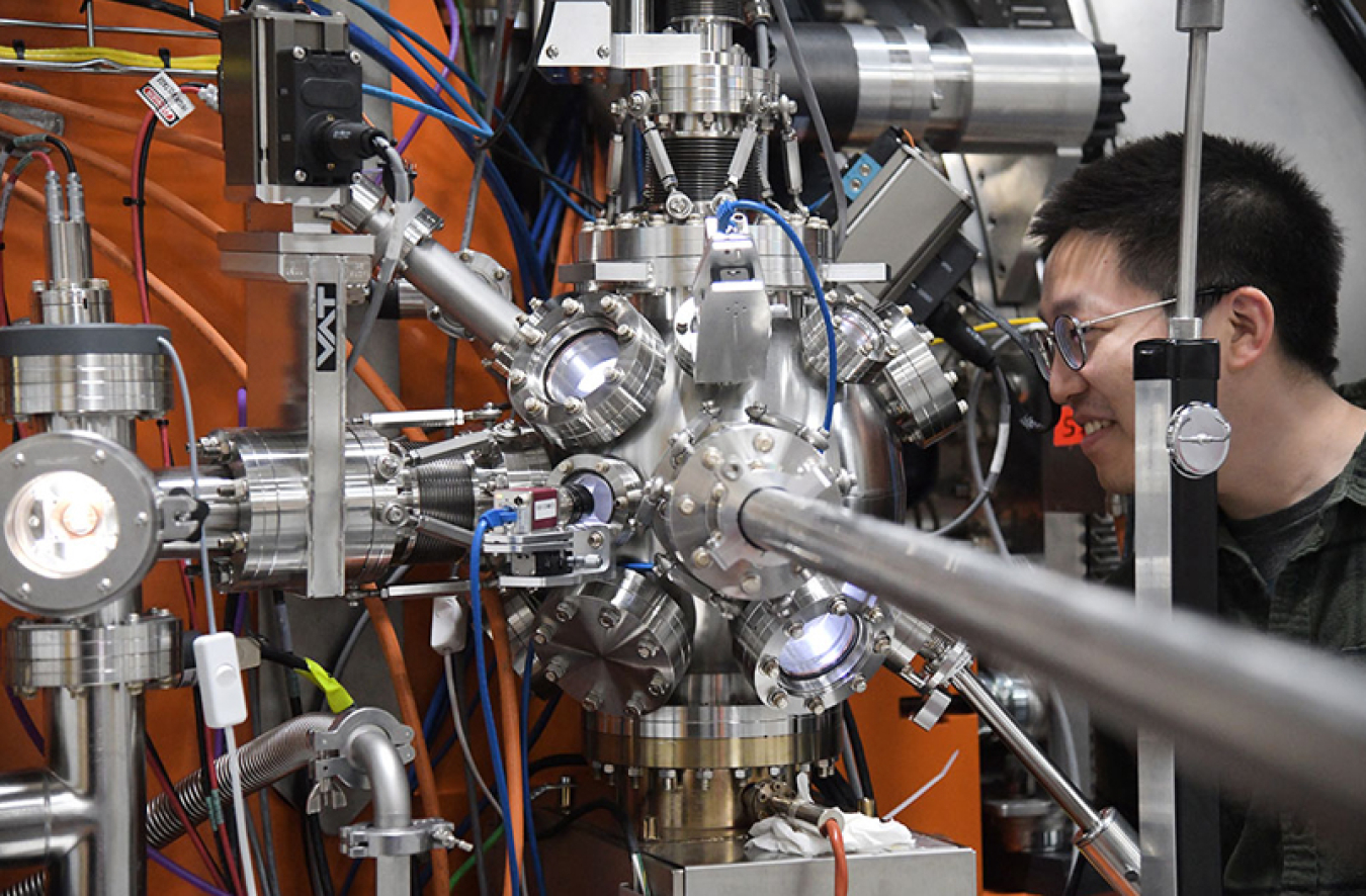 Yao Shen looks at a section of the SIX beamline of the National Synchrotron Light Source II (NSLS-II). The section of the machine has a silver-colored sphere with various lenses on the outside of it and a pole sticking out of it towards the camera. This section of the machine is for transferring the samples into the sample chamber.