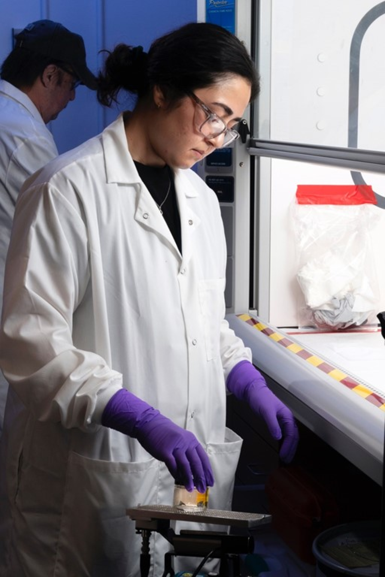 A woman dressed in a lab coat and rubber gloves prepares to put a forensic sample into a machine.