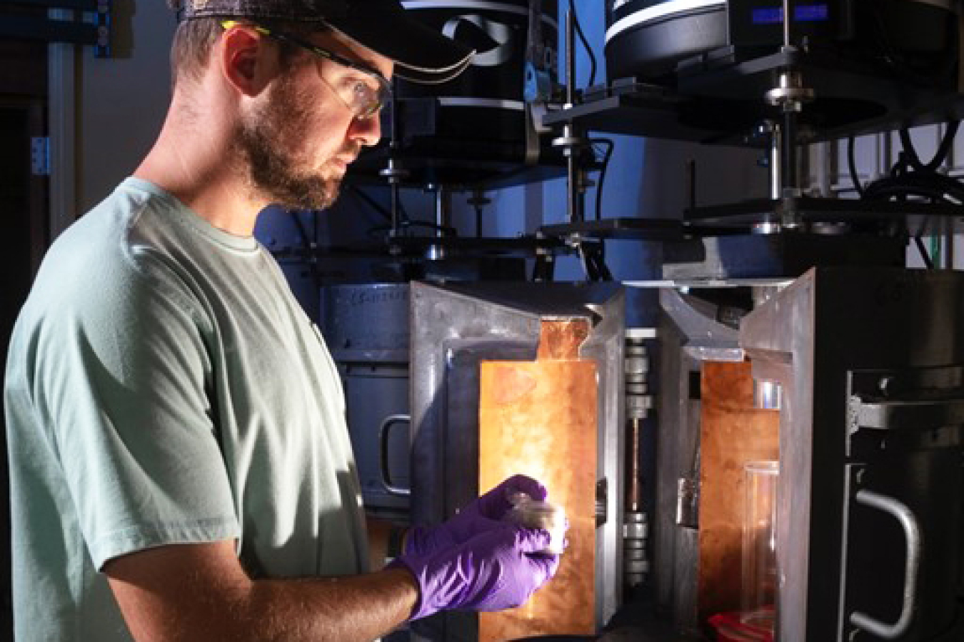 A man in stands in front of a lab machine with its door open. He is preparing to put a sample inside. The open door casts a glow upon his face.