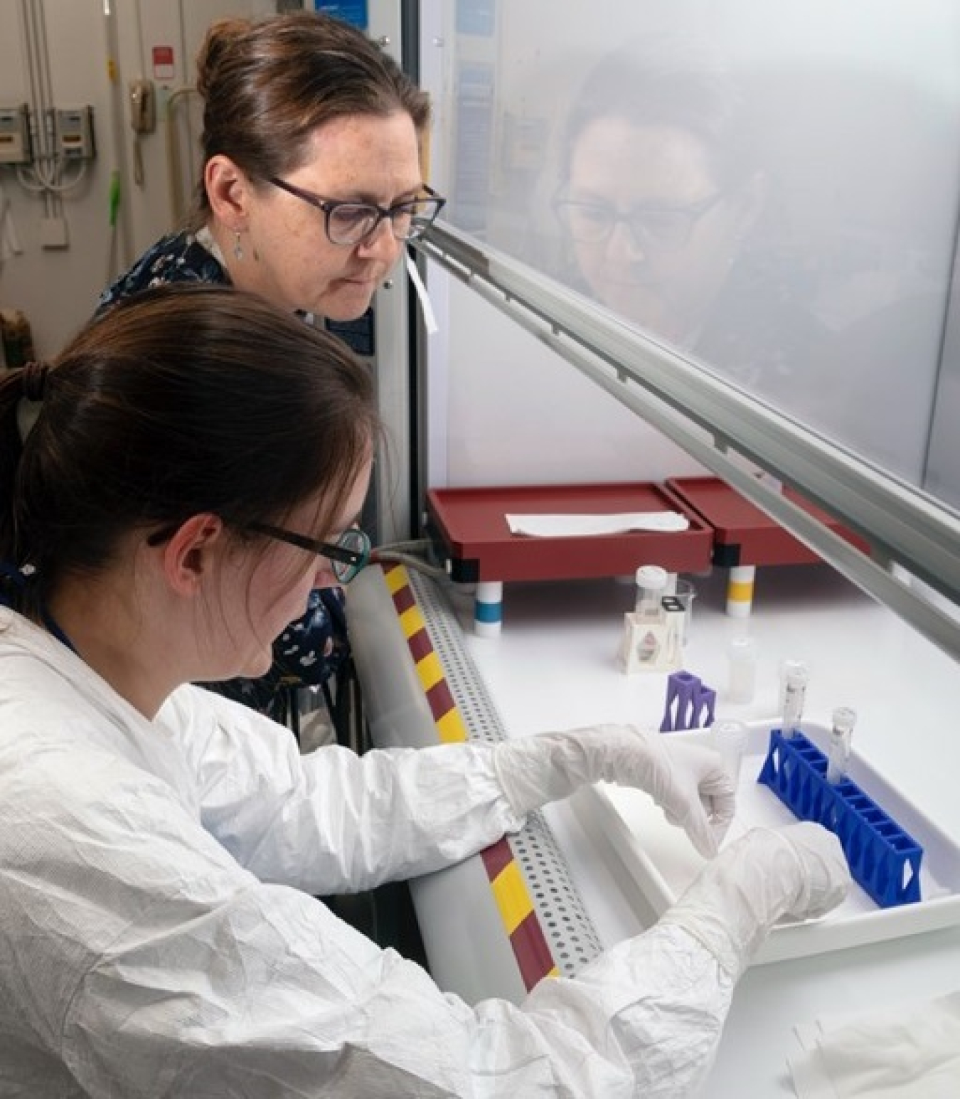 Two women dressed in white lab coats and purple rubber gloves concentrate on a sample one of them is holding. They are preparing to put it into a machine.