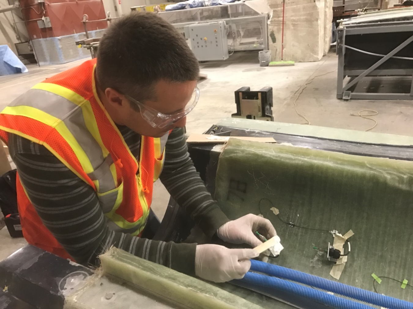 A man with safety goggles and a safety vest works on a wind turbine blade in a warehouse environment.