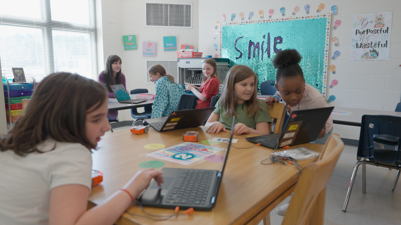 Students in a classroom sit at tables and work on laptops while talking and laughing with each other