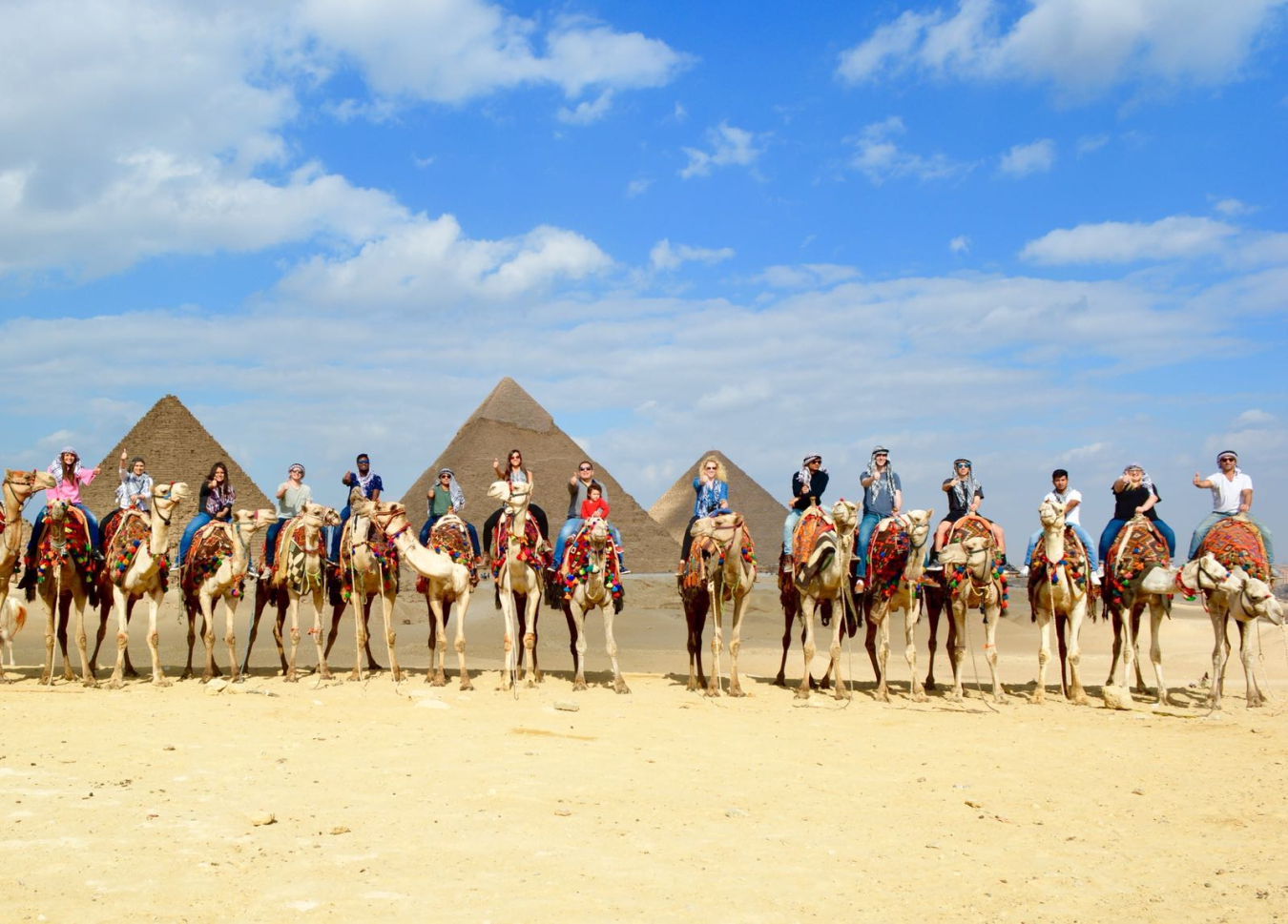 Fifteen people sitting on camels in front of pyramids