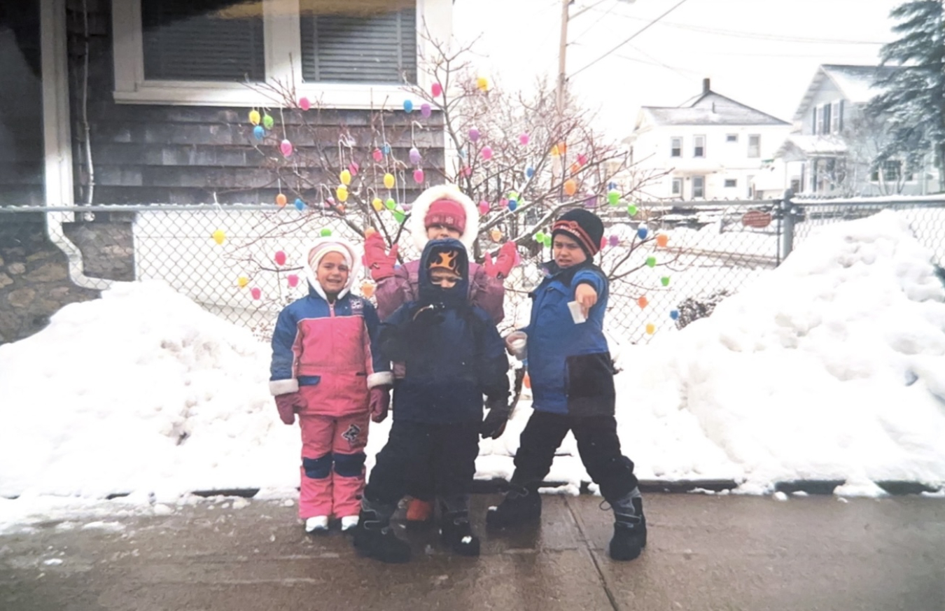 Four kids dressed in winter gear standing in front of a snowy yard.