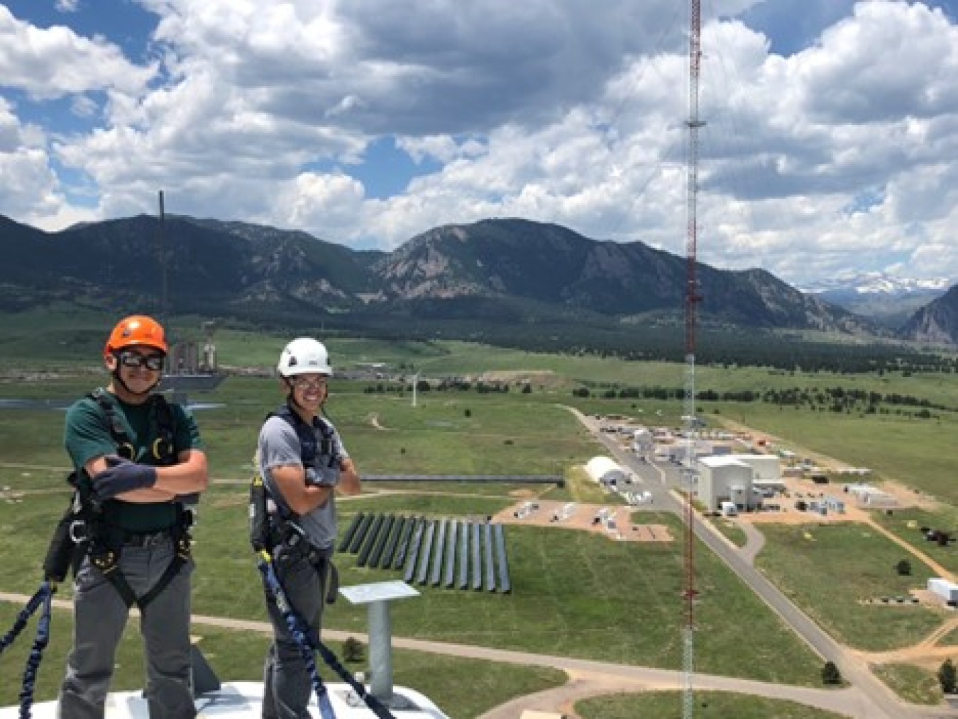 Andrew Simms and a coworker wear helmets and climbing gear while standing on top of a wind turbine