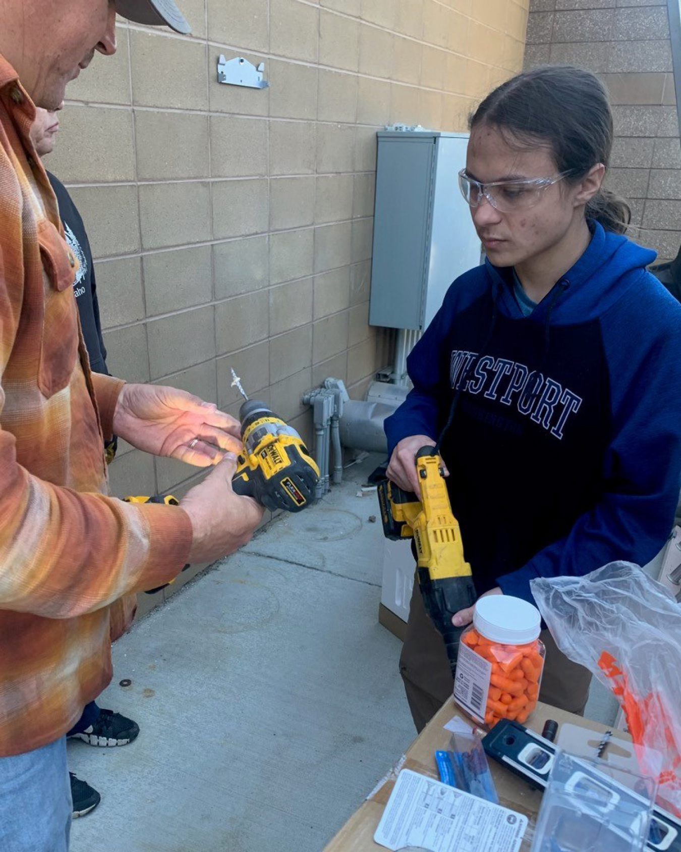 A man shows a younger man power tool features. A jar of something orange, which could be cheese puffs or electrical connectors, is on the table in the foreground.