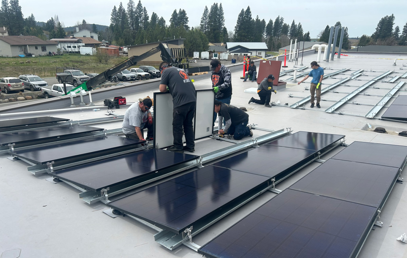 Workers install rooftop solar panels with pine trees in background.