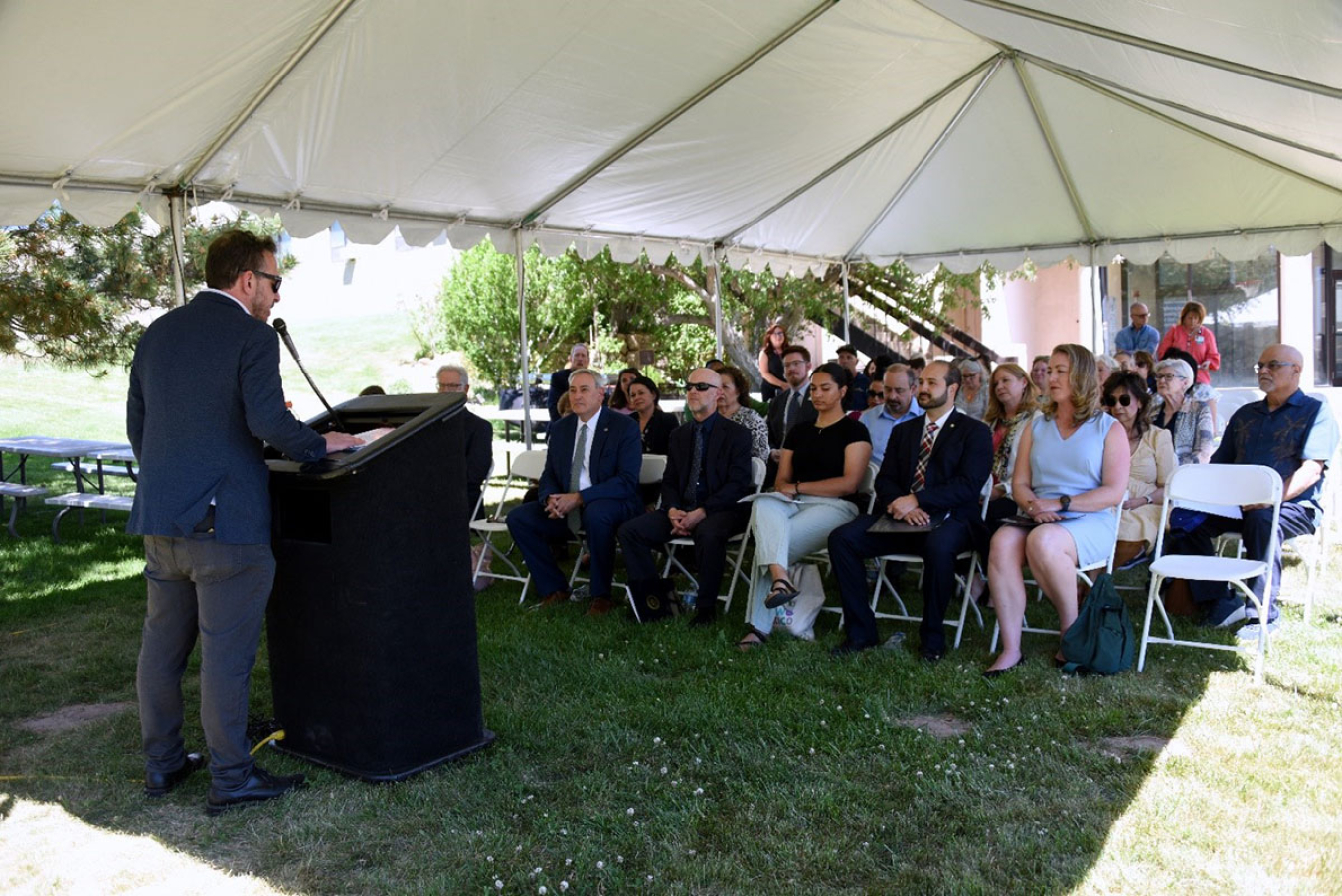 Jonathan Corbin, PhD, Co-Director, Worker Health Protection Program speaks to the former workers from a podium. This is under a tent on a lawn.