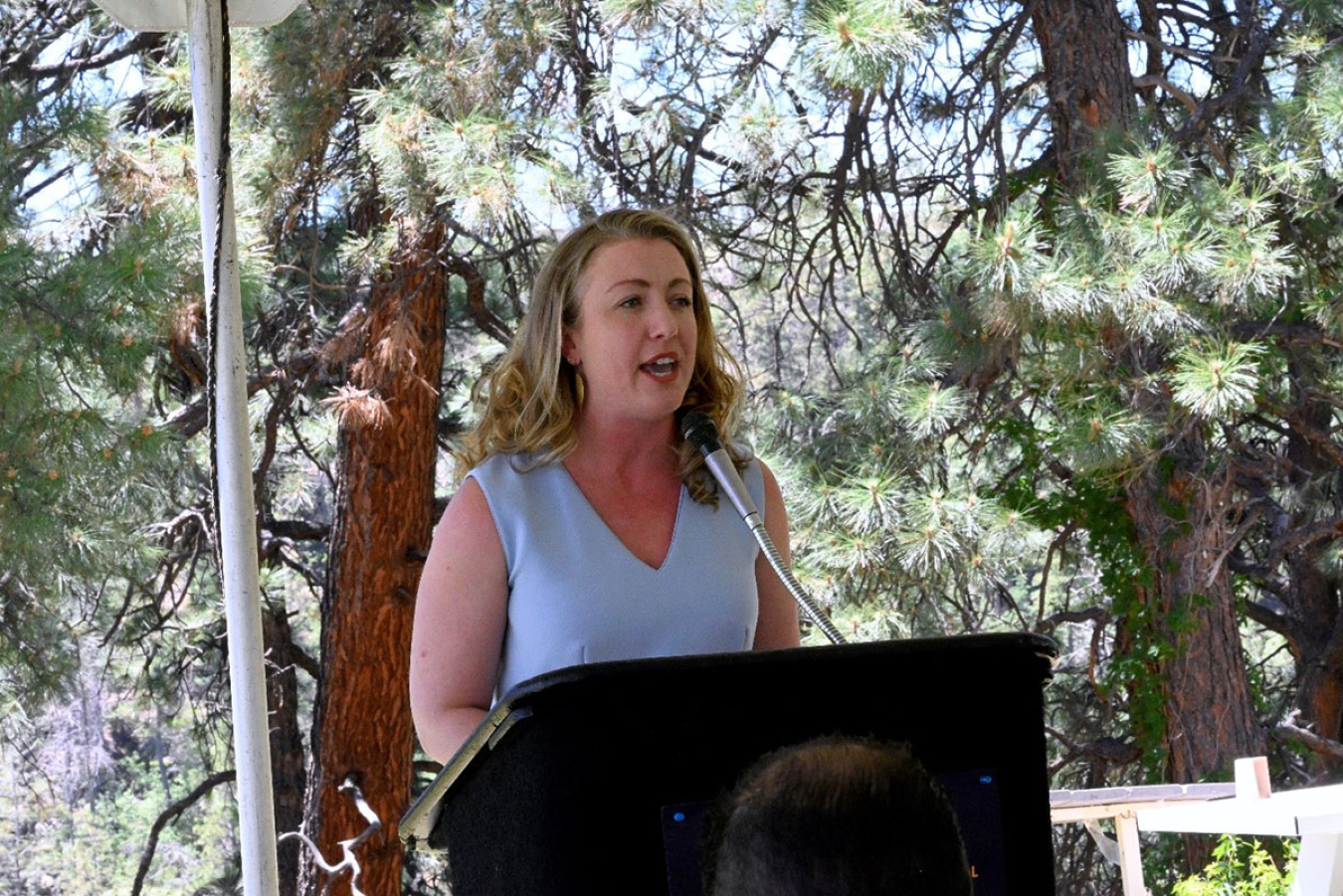 Rita O’Connell, Office of U.S. Senator Heinrich. speaks to former workers from the podium. This is under a tent with trees in the background.