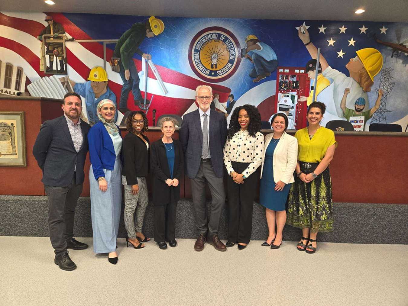 (Left to Right) CUNY: Jonathan Corbin, PhD; Khaula Khatlani, MD, MSc; Tiffany Martin, MA; Amy Manowitz, MPH; Steven Markowitz, MD, DrPH; Amaka Onyekelu-Eze; and JHU: Aisha Rivera, MD, MS; and Joslyn Cordova pose for a photo. This is done in front of a mural for the International Brotherhood of Electrical Workers.