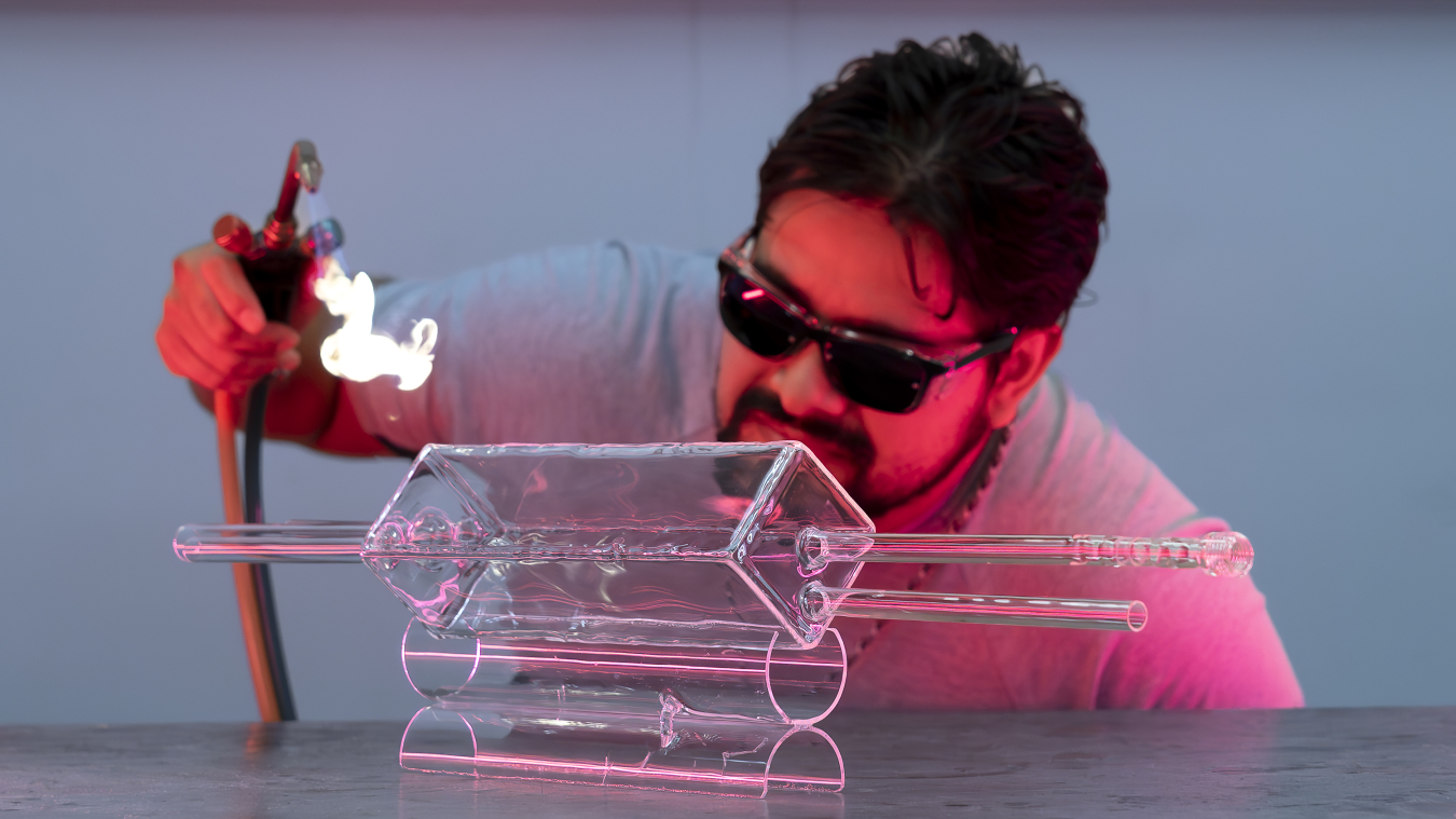 man wearing sunglass standing being clear glass rectangle with small blowtorch in his right hand