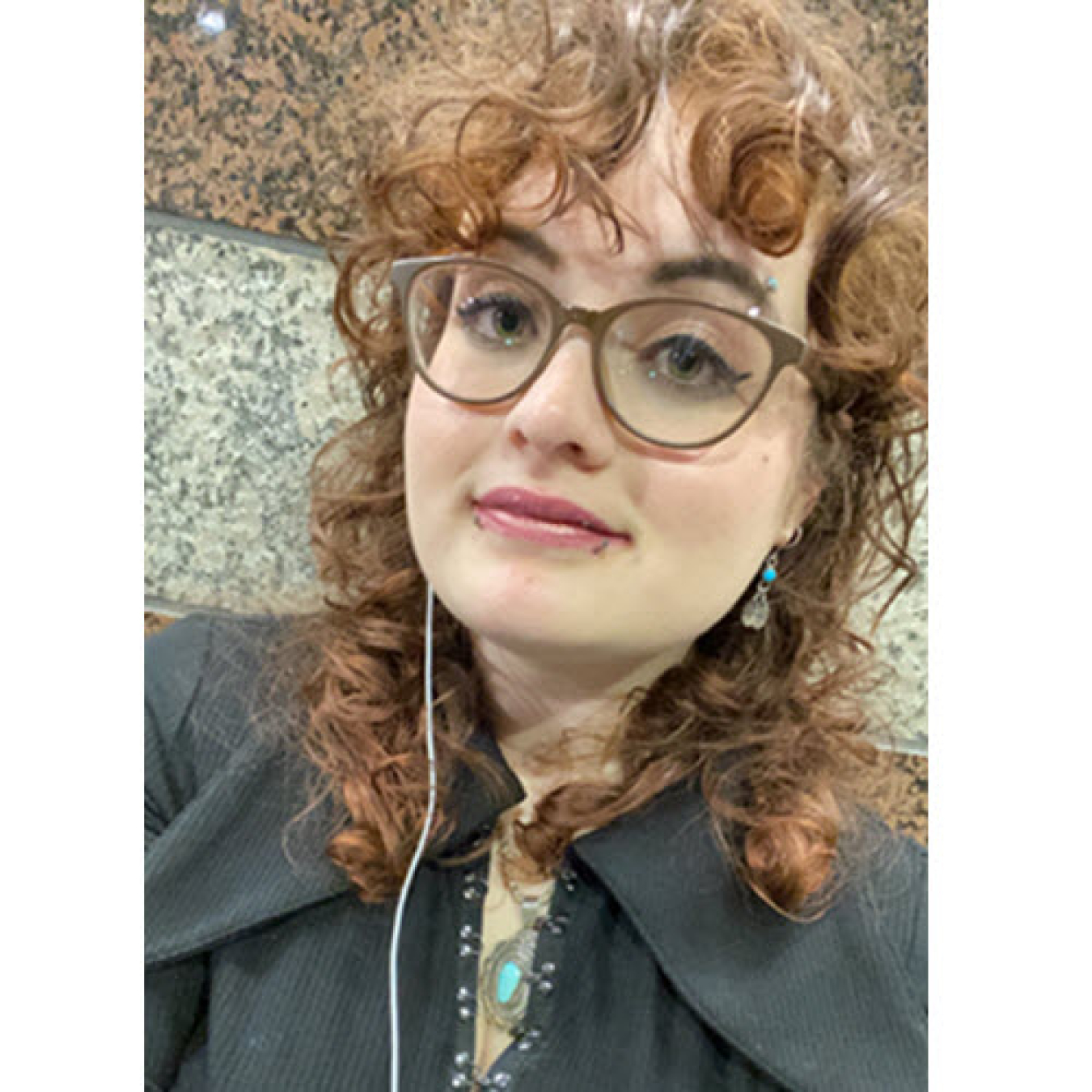 Headshot of Alexis Glaudin, a young woman with curly hair wearing glasses and wired earbuds.