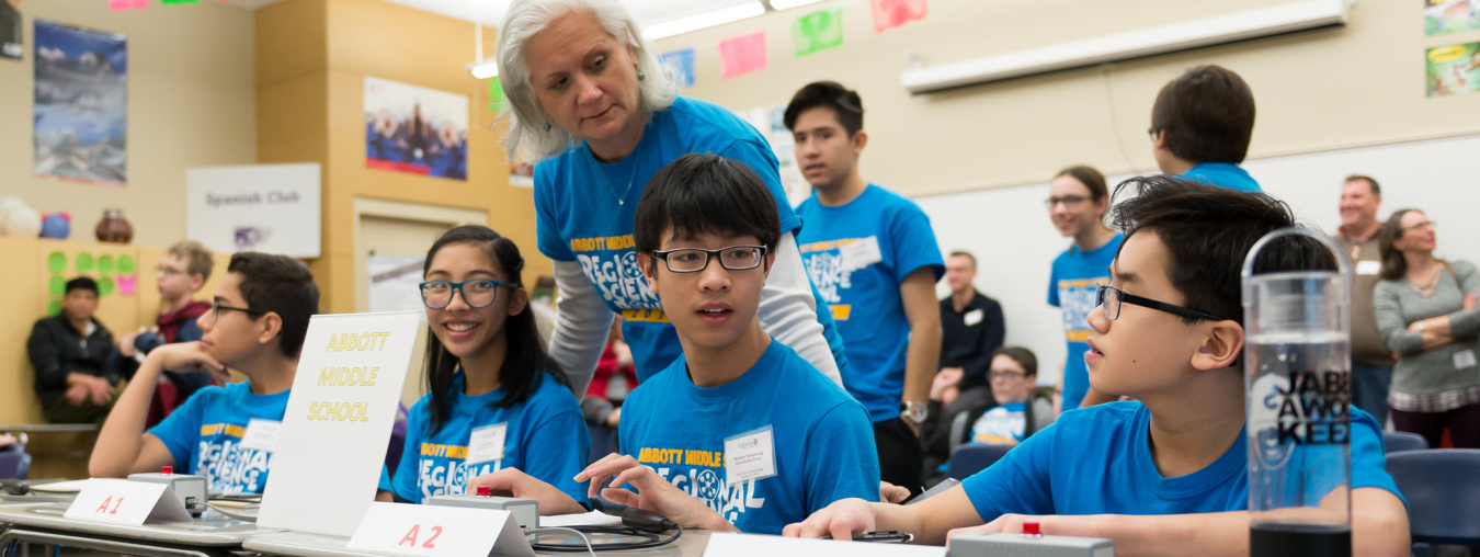 Students wearing matching blue t-shirts sit at a table with buzzer buttons. A mentor stands behind the students, glancing at them.