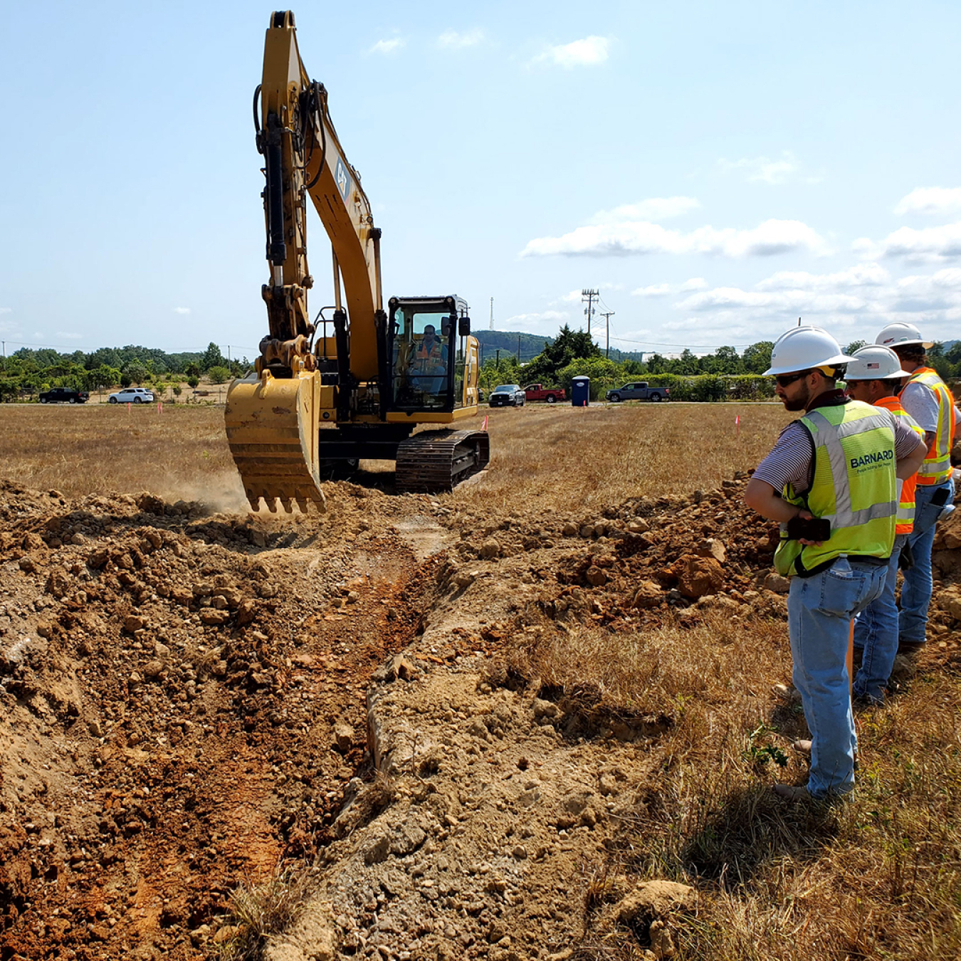 Yellow excavator in field, three people in hard hats watch