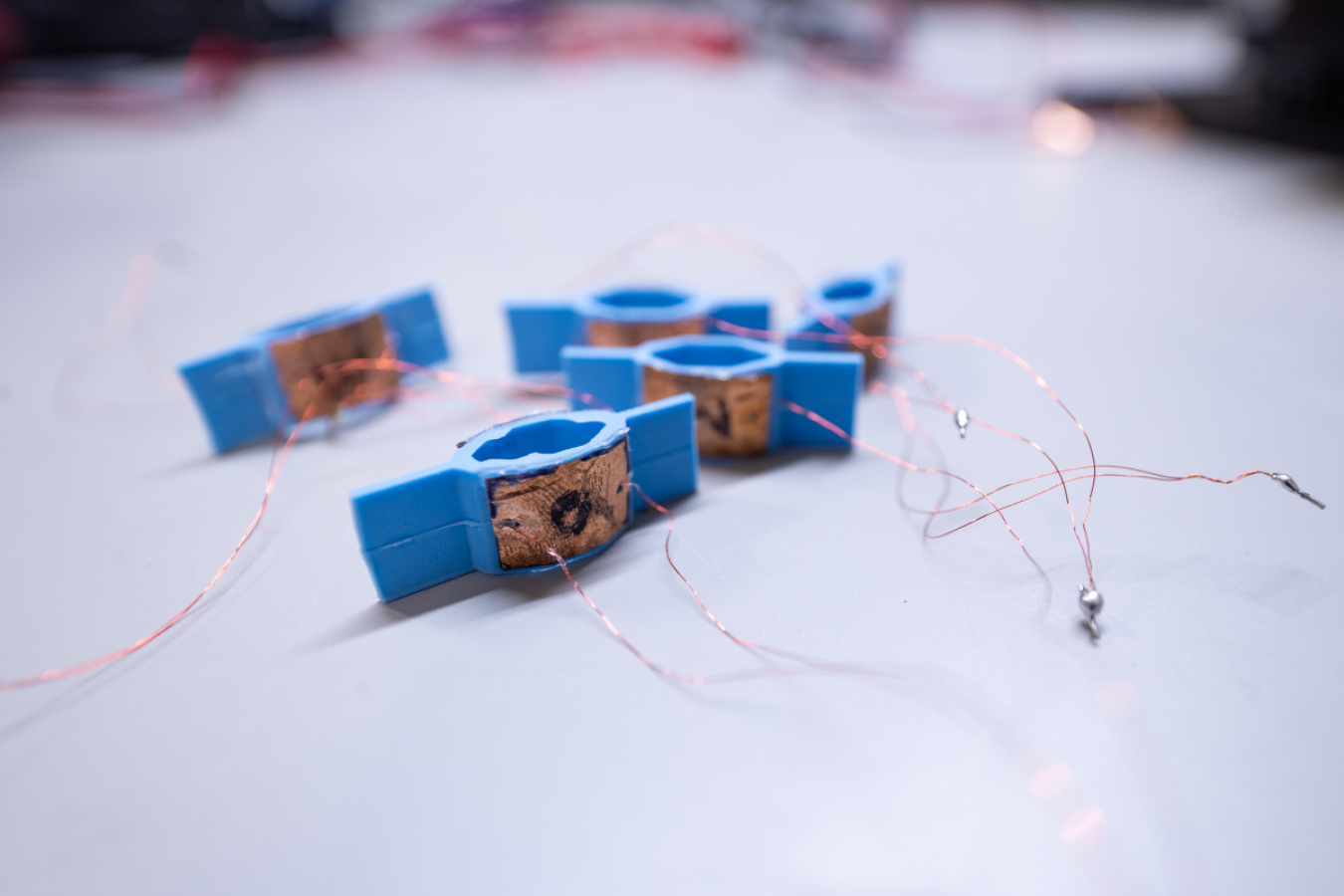 Five blue devices with oval centers and flat pieces positioned on the top and bottom side of the oval sit on a flat surface. Wires are coming off of each device.