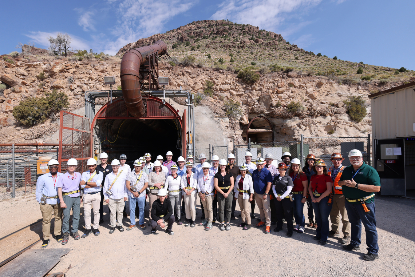 A large group of people wearing hard hats stands in front of a mine tunnel. Railroad tracks are to the left.