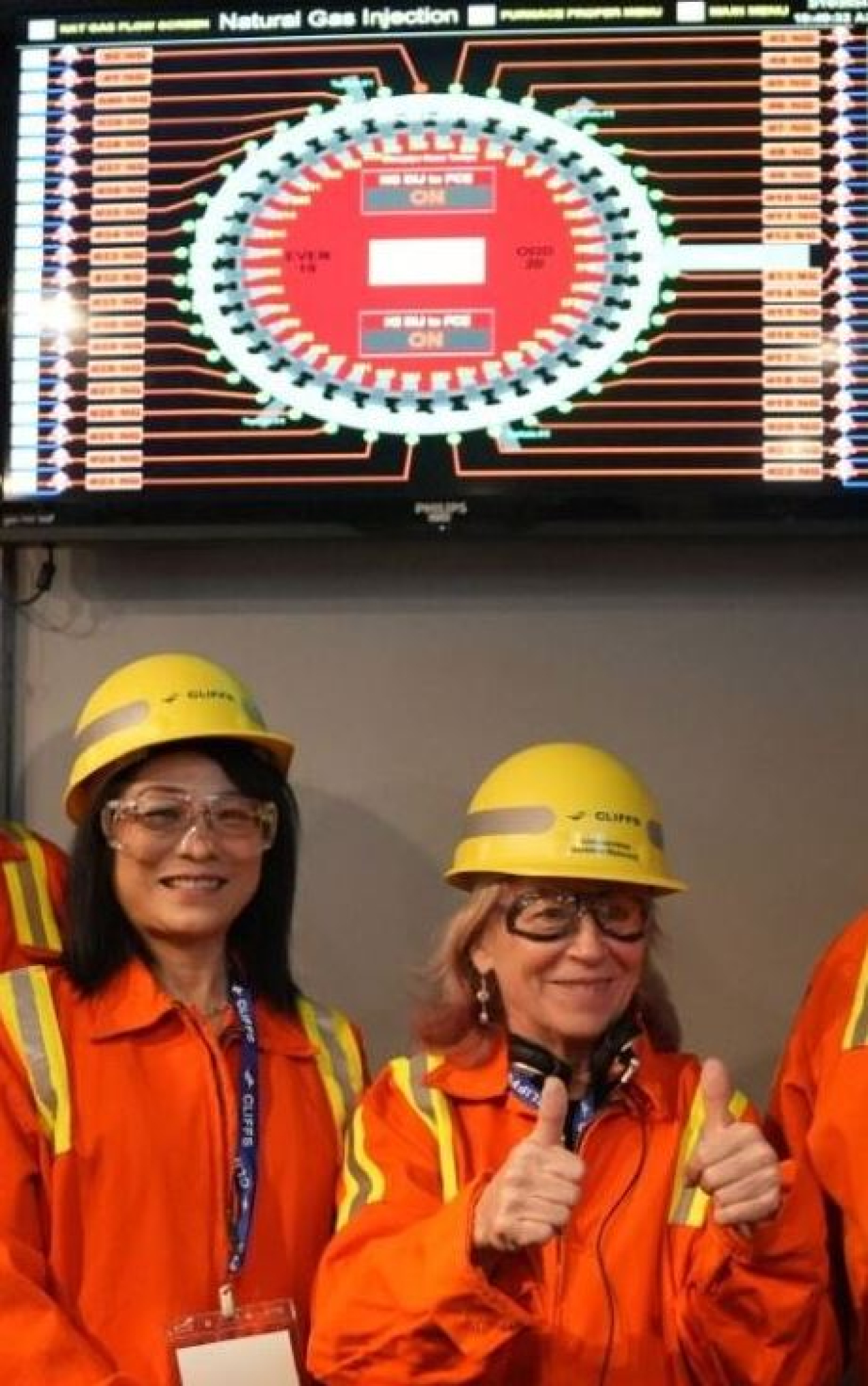 Two people wearing protective gear standing underneath a monitor displaying plant system status