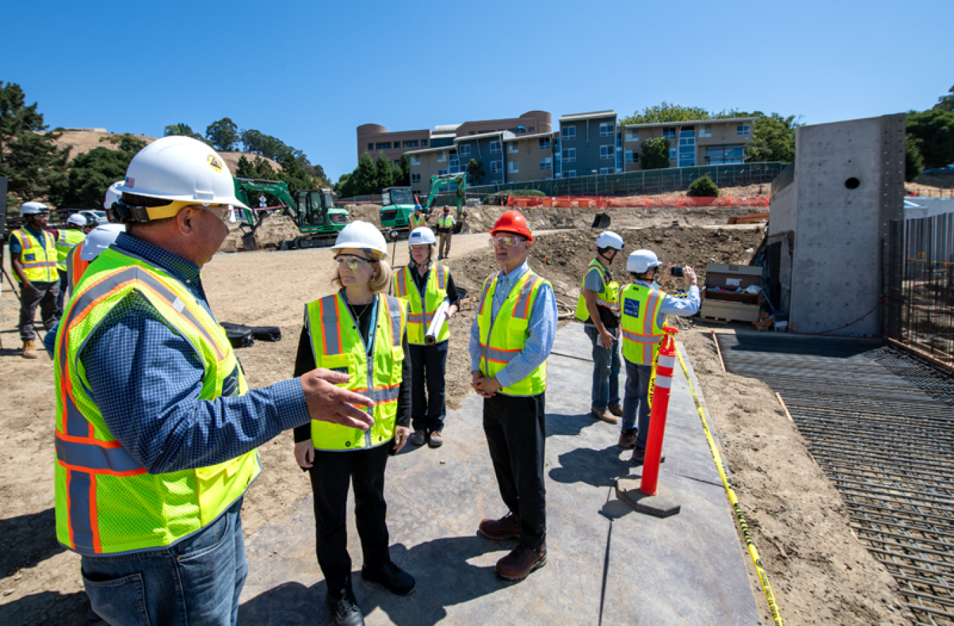 A number of Berkeley Lab’s leaders in construction equipment, including hard hats and safety vests. They are standing on a concrete pad, with ground broken for construction to the right of them.