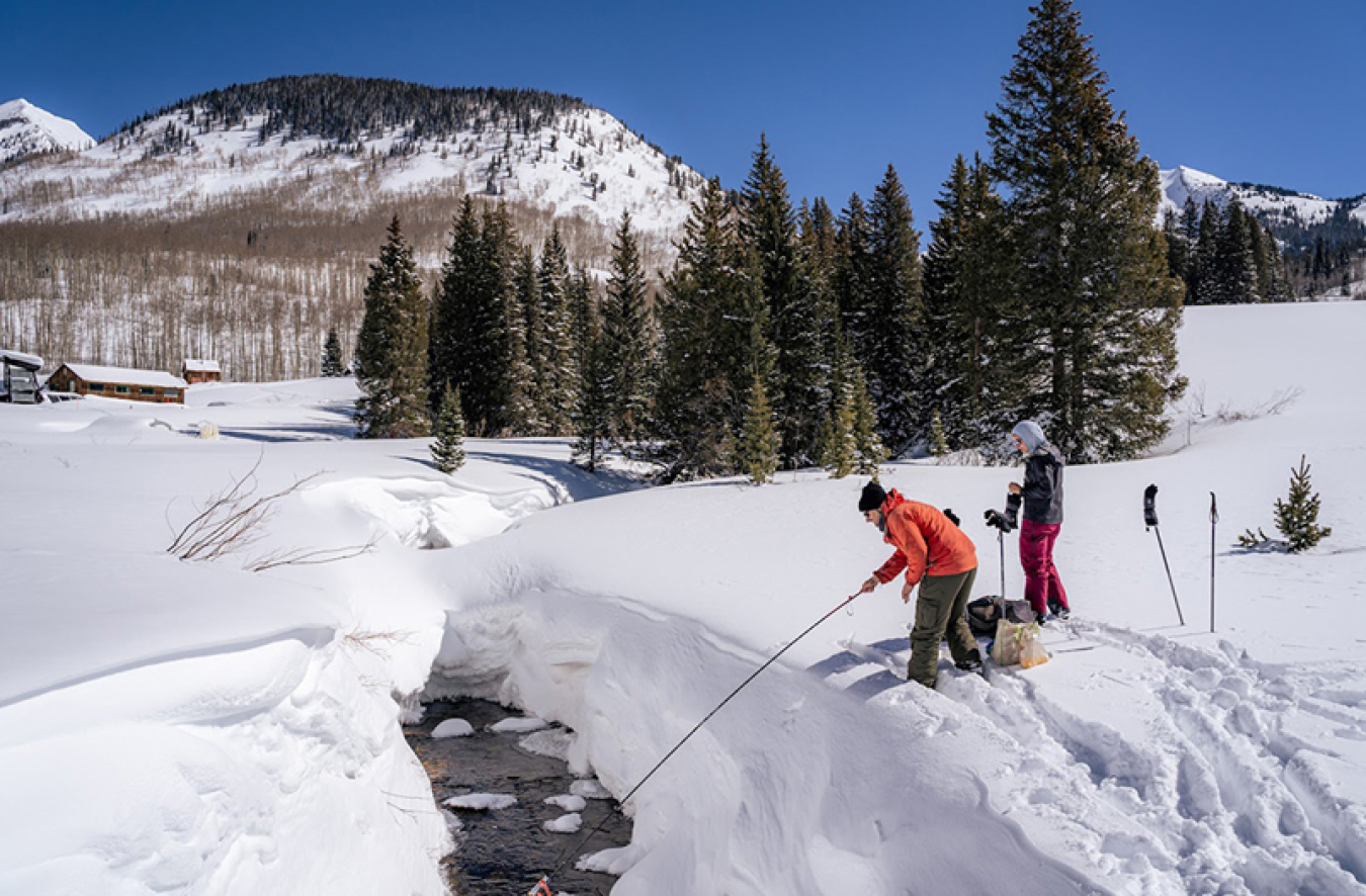 Two scientists in cold-weather clothing are standing in a snowy area next to a stream with mountains in the background. The one on the left is sticking a probe in the stream. There are hiking poles stuck in the snow behind both of them.