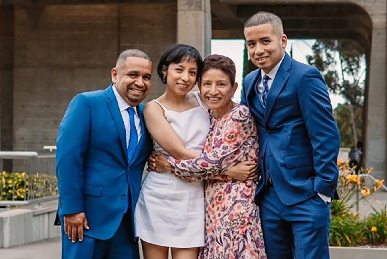 Four people dressed formally, including Gabriela Quintanilla, pose in a blooming garden with a large building behind them.