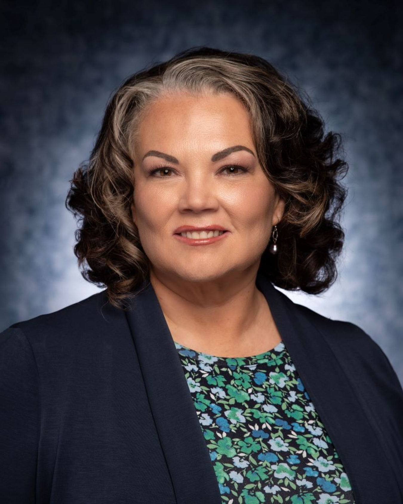 A headshot of a woman with shoulder length hair, a floral shirt, and a black blazer