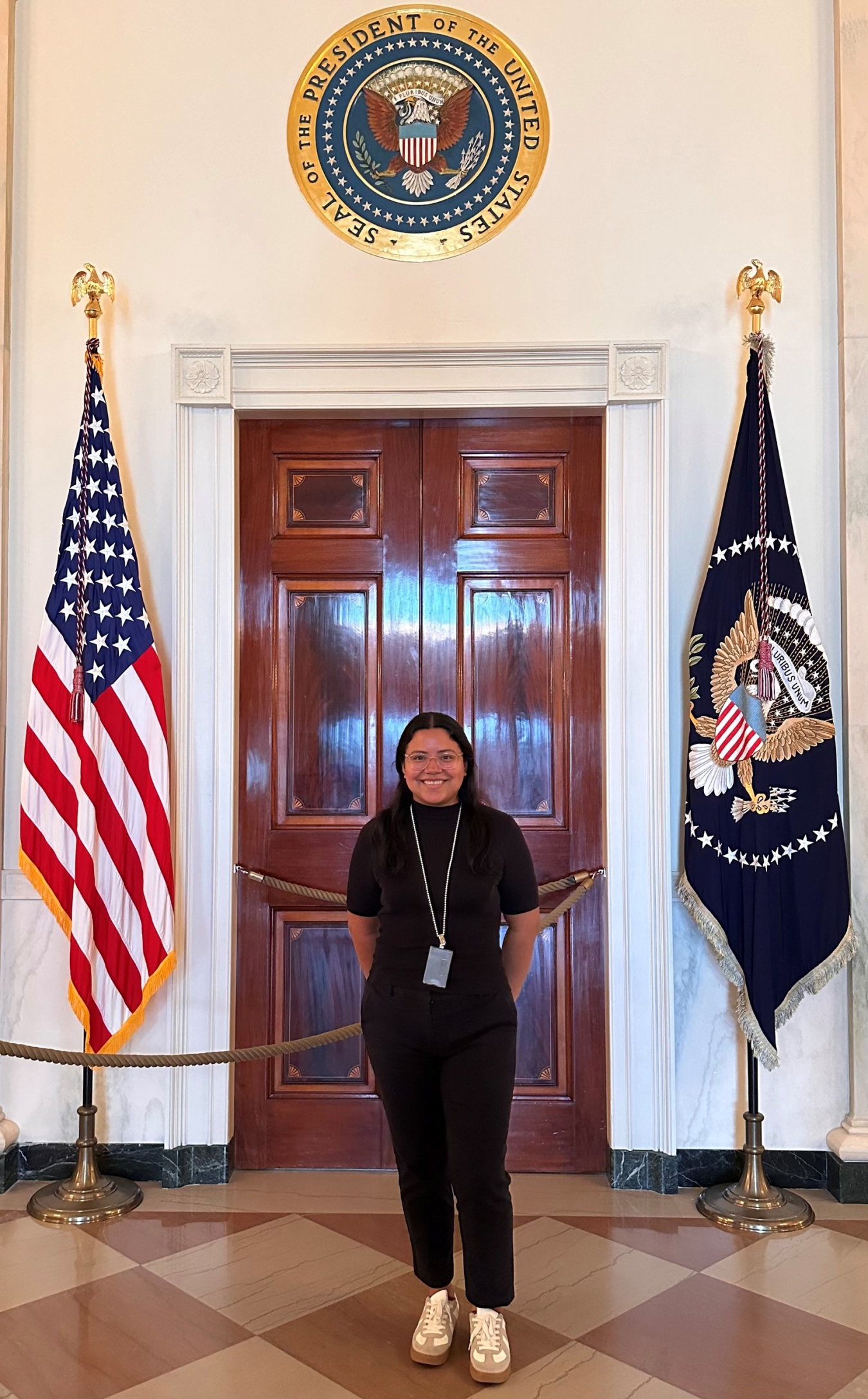 Cecilia Villegas standing in front of wooden door with American flag on one side and Flag of the President of the United States on the other side.