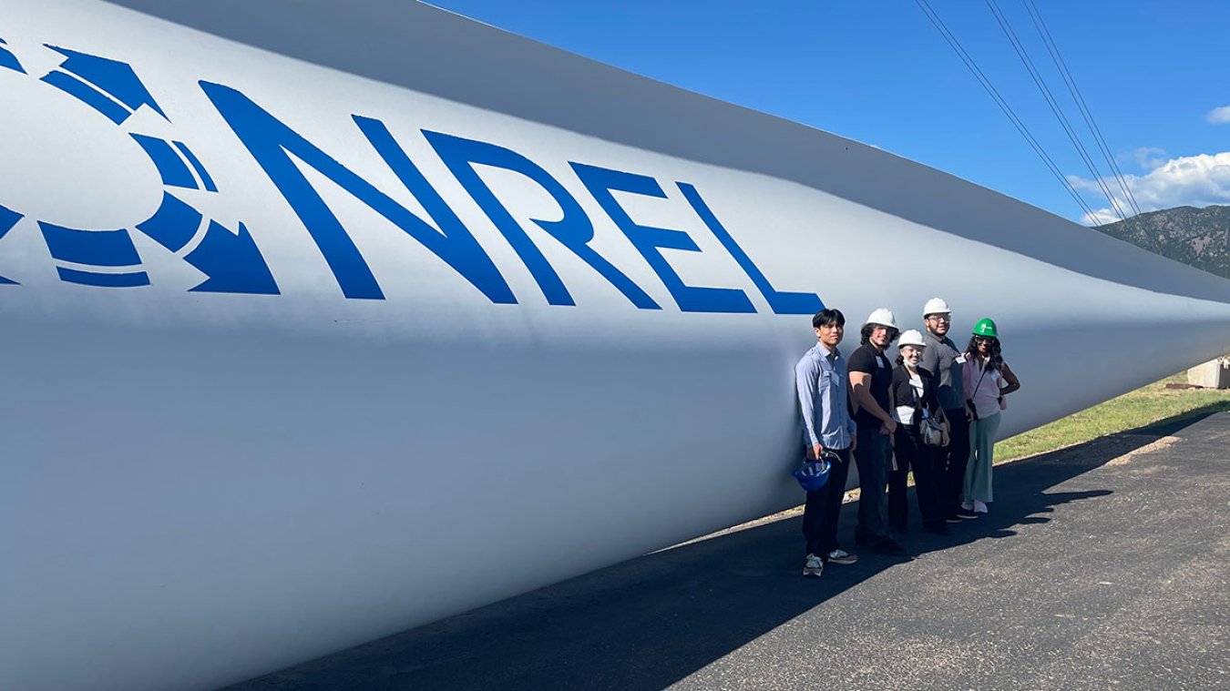 Four indiviuals wearing hard hats standing in front of the blad of a wind turbine. The blade has the logo for the National Renewable Energy Laboratory on its side.