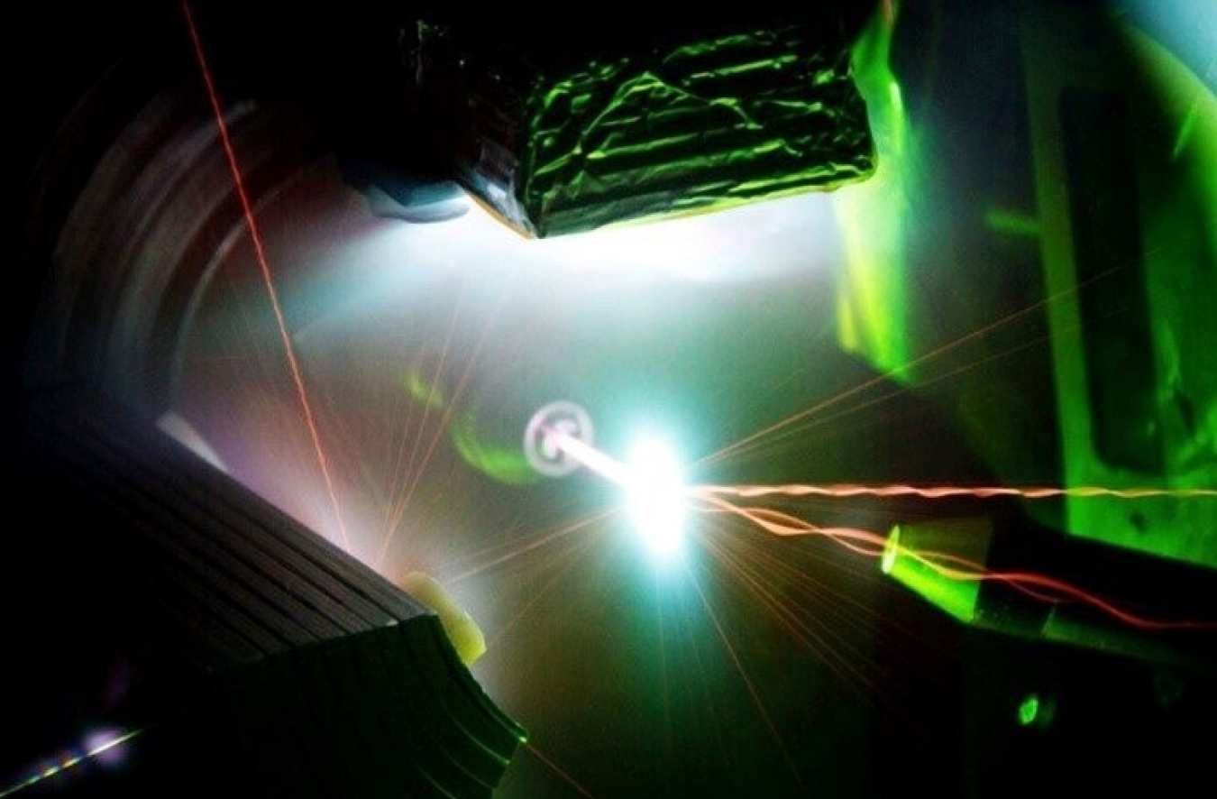 The image is a dark background with a bright white light at the center, showing the interaction of invisible infrared light from the 200-trillion-watt Trident Laser with a one-micrometer thick foil target in the center of the image, generating a high energy density plasma. The interaction that is occurring is lime green and white; the background of the image is black.