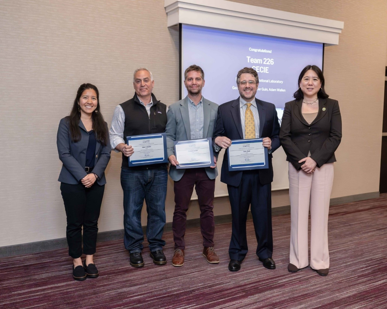 A group of five people pose and smile for the camera, three are holding certificates. 