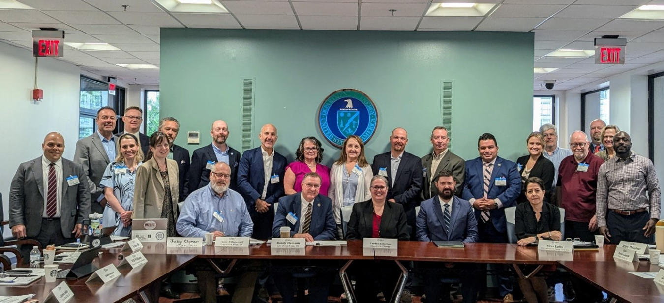 A large group of people in a conference sit and stand for a group photo. The DOE seal is hung on the wall behind them.