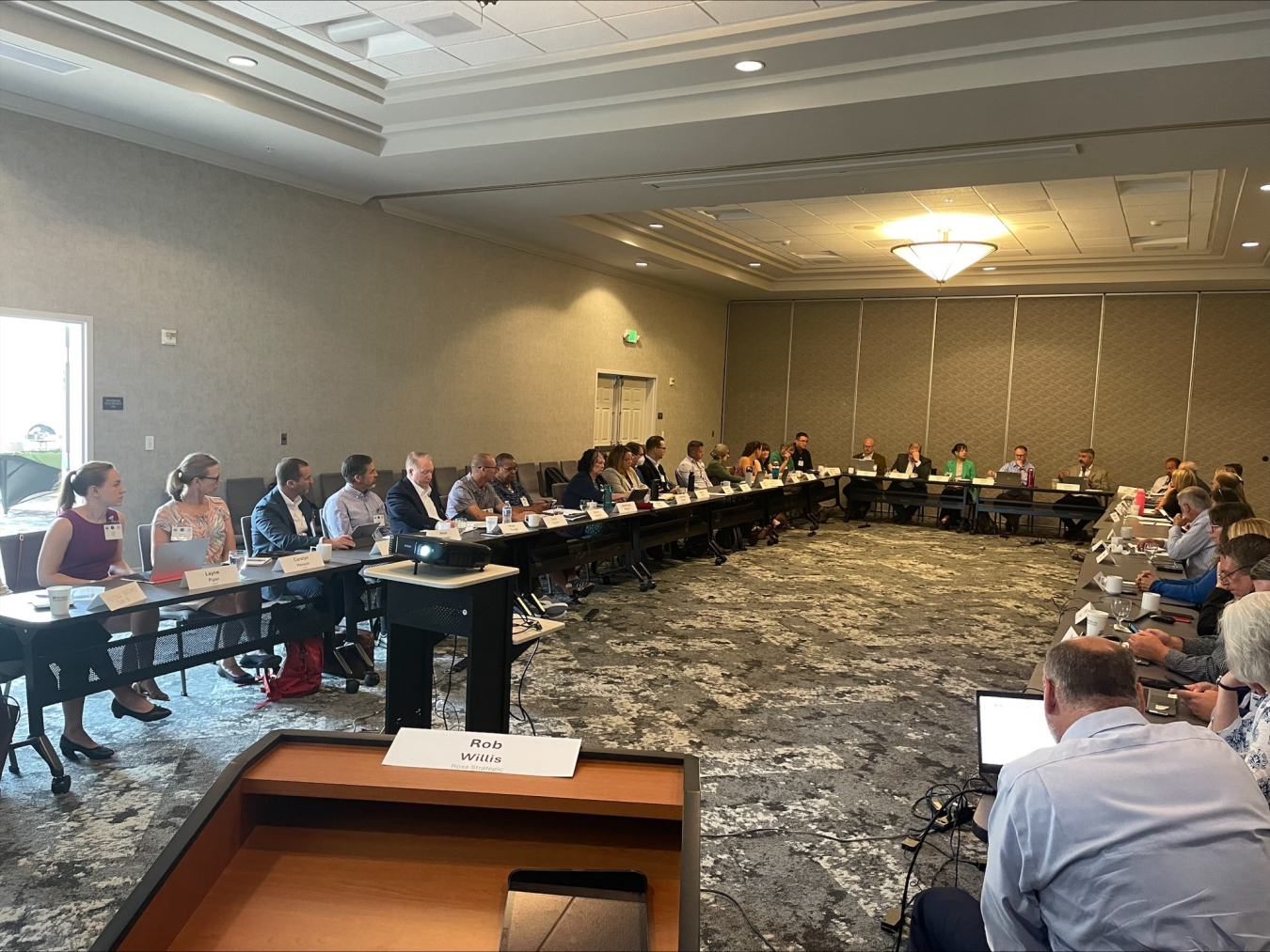 A large conference room with tables along the wall and people sitting at the all the seats around the tables