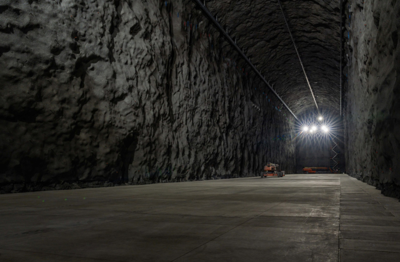 A long stone tunnel with a concrete floor. There are bright lights at the end of it.