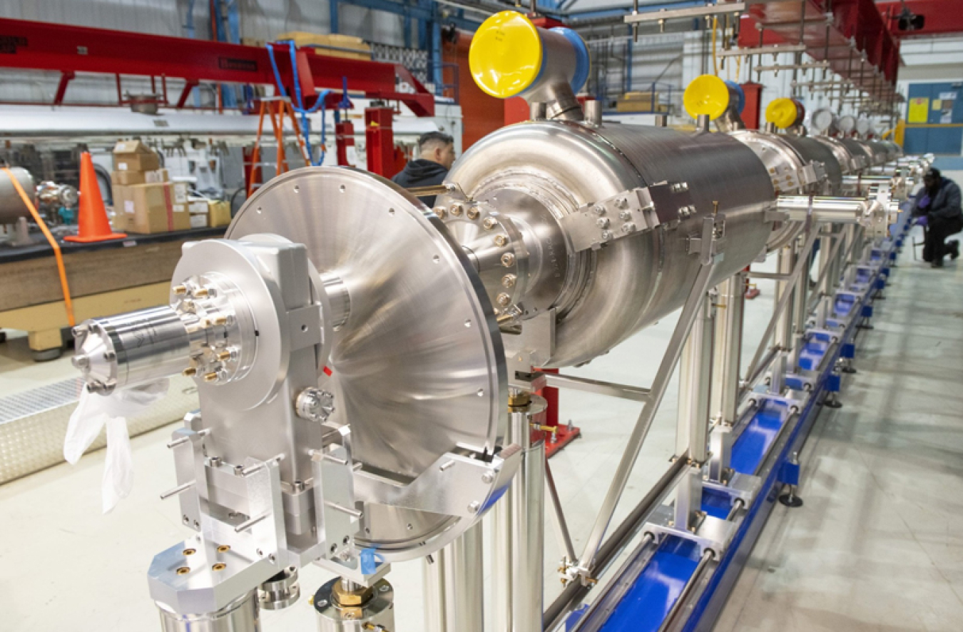 A long view of the fully assembled prototype high-beta 650-megahertz cryomodule shows a glossy steel horizontal structure that is large and heavy. It is in a lab area where it is being built out. Four of these will make up the final stage in Fermilab’s new linear accelerator.