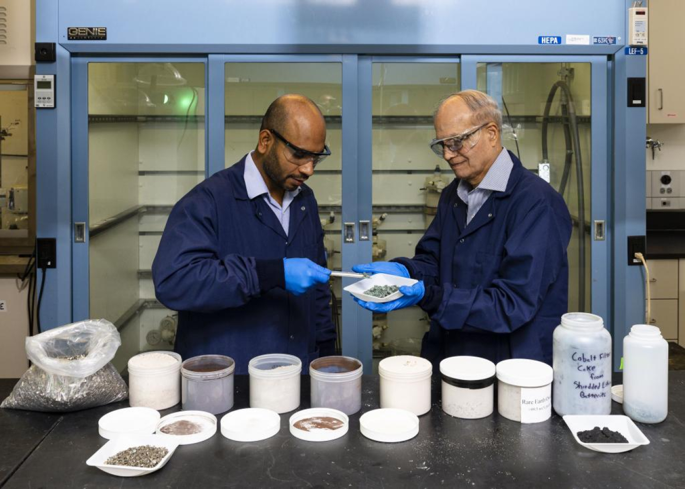 Two scientists in protective gear look at samples in several pots in a lab