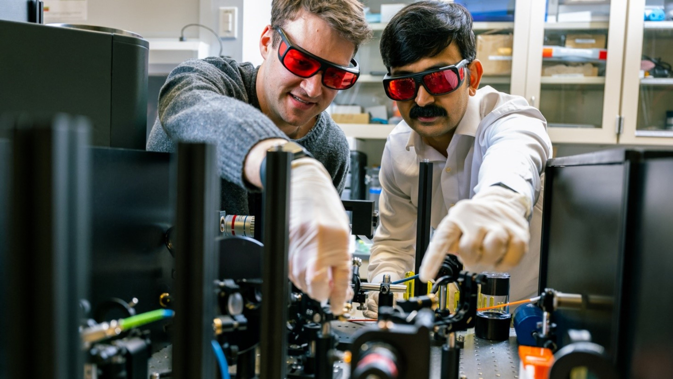  Two men in safety glasses work on a piece of equipment in a lab setting. 