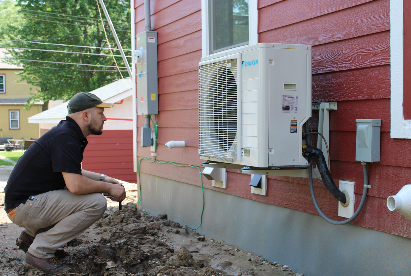 A crouching person inspects an air-source heat pump mounted to the side of a home.