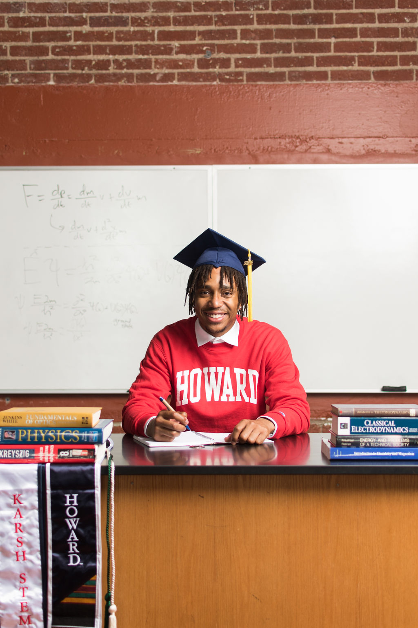 Jon-Edward Stokes wearing a Howard University sweatshirt and graduation cap while sitting at a desk with books 